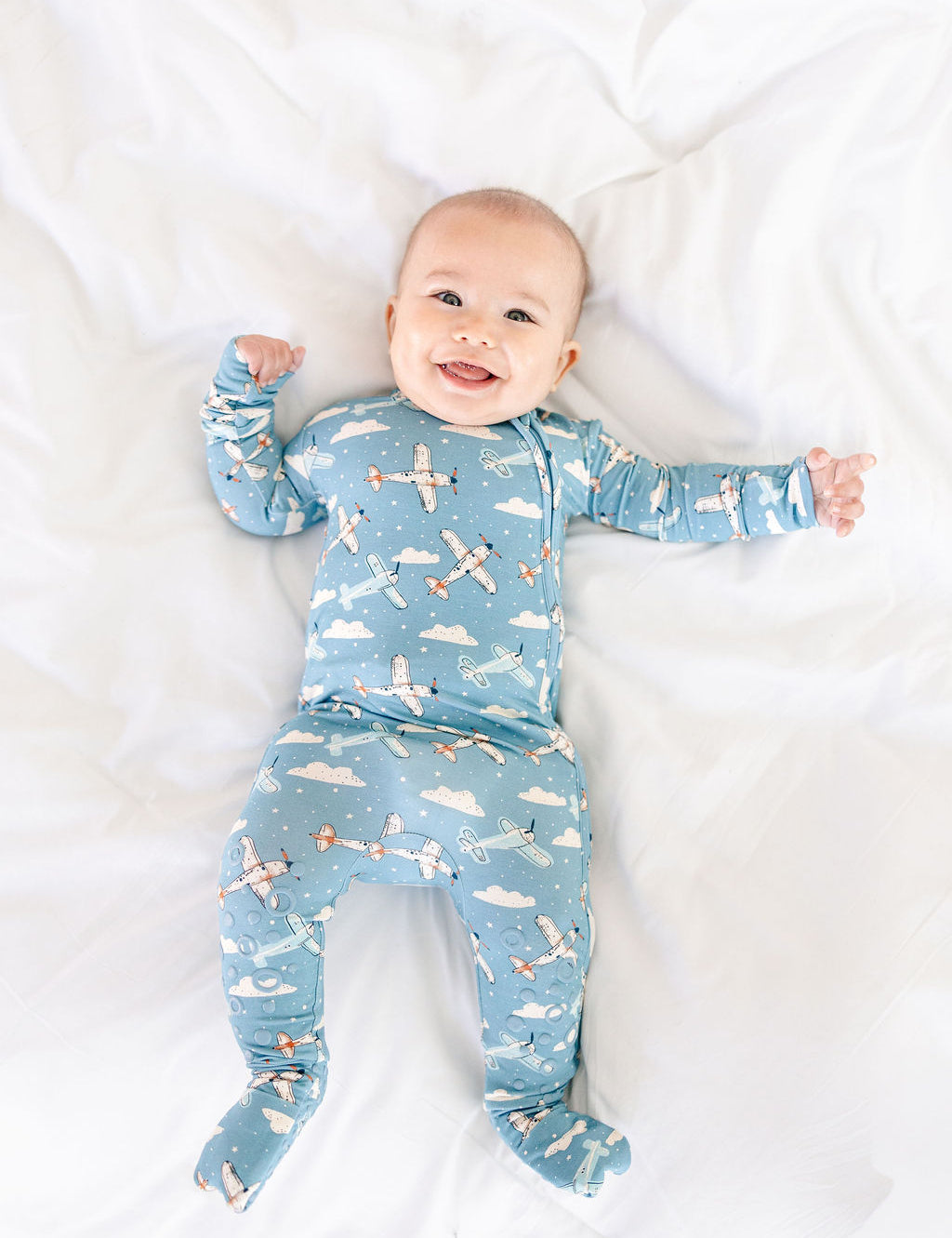 Baby wearing a blue airplane and cloud patterned onesie on a white blanket