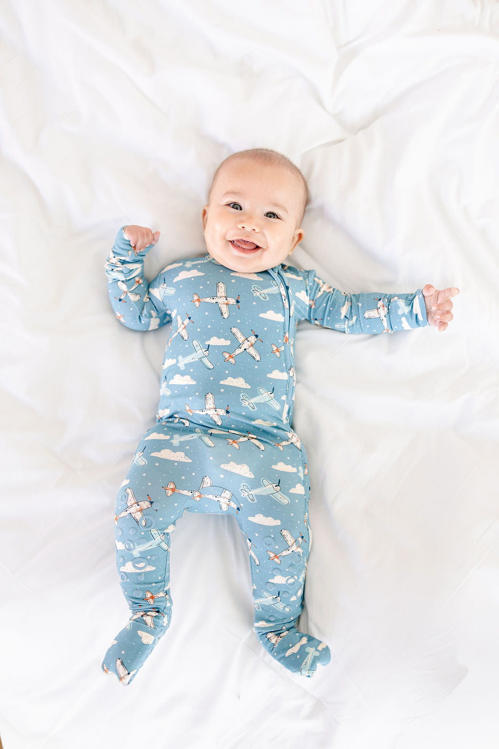 Baby wearing a blue airplane and cloud patterned onesie on a white blanket